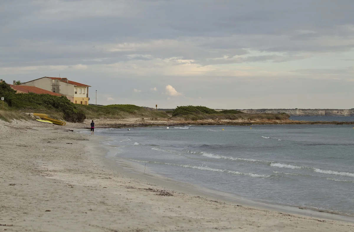 Sandy shoreline at Putzu Idu on Sardinia's San Vero Milis coast, with shallow waves, small yellow boats, coastal houses and distant cliffs.